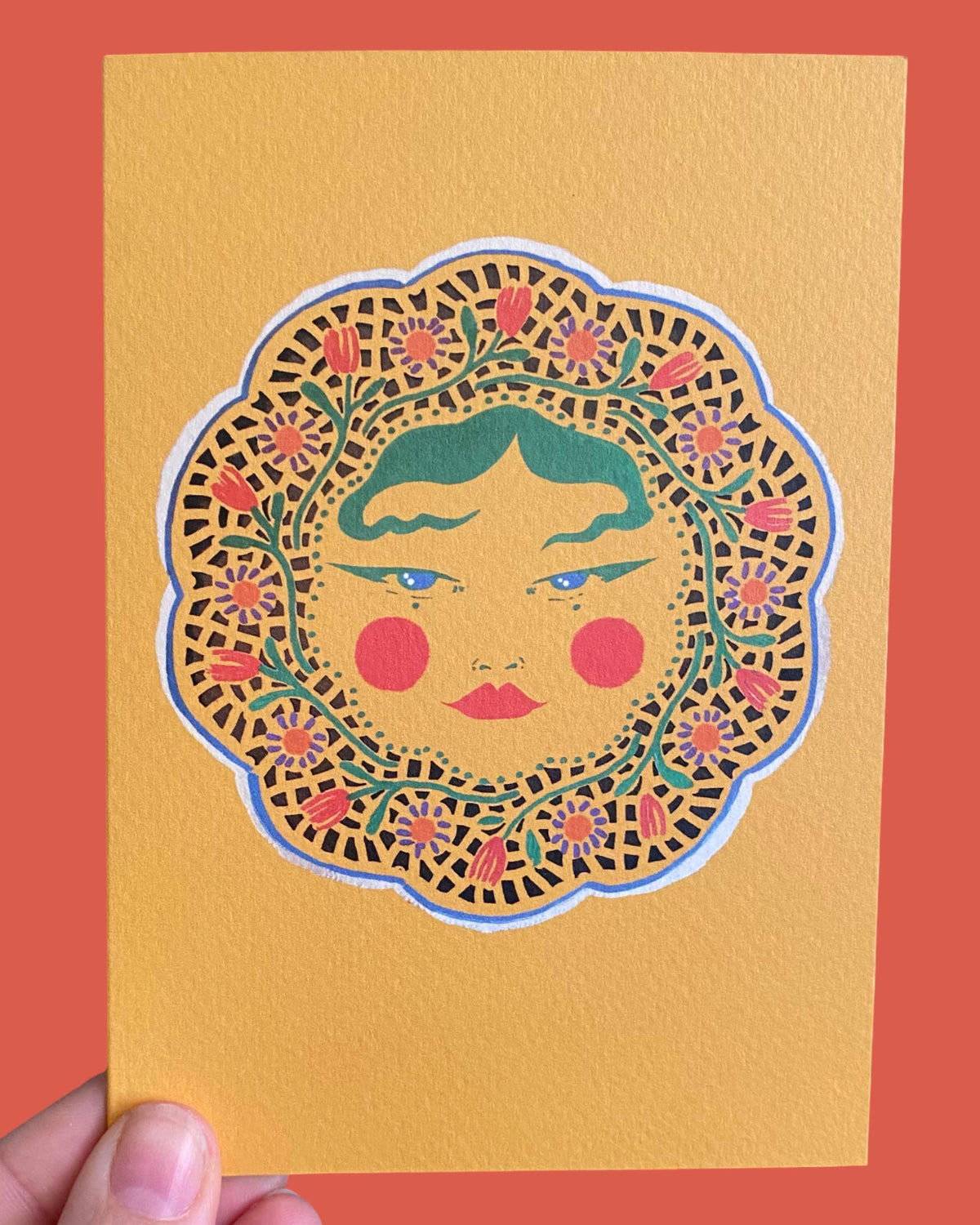 A hand holds a textured mustard yellow art print or card featuring a stylized face framed by a floral mandala border, set against a solid coral-red background.