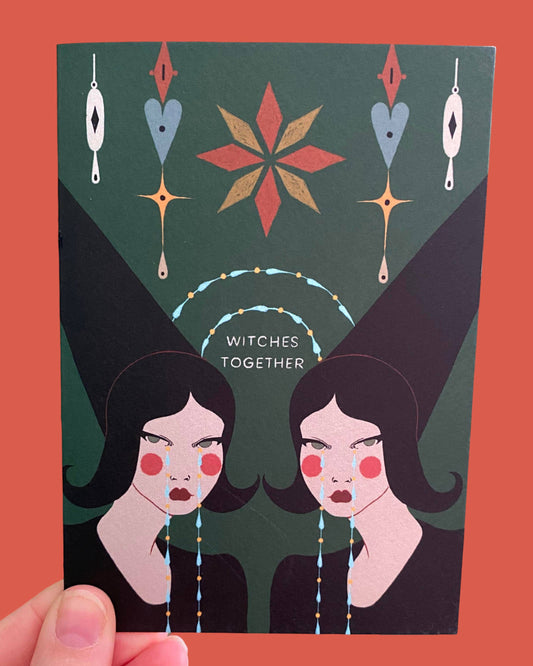 A hand holds a square art card featuring a stylised illustration of two women with pointed hats and the text 'WITCHES TOGETHER' on a dark green background, set against a solid coral-red backdrop.