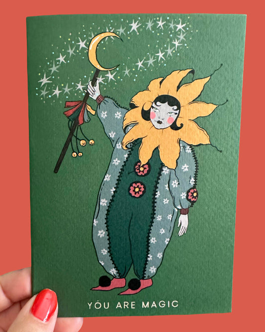 A hand holds a green illustrated card featuring a whimsical sunflower-like character with a crescent moon and stars above, with the text 'YOU ARE MAGIC' at the bottom, against a coral-red background.
