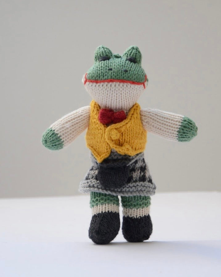 A hand-knitted frog doll wearing a yellow vest, red bow tie, and a dark patterned skirt, standing against a plain light grey background.