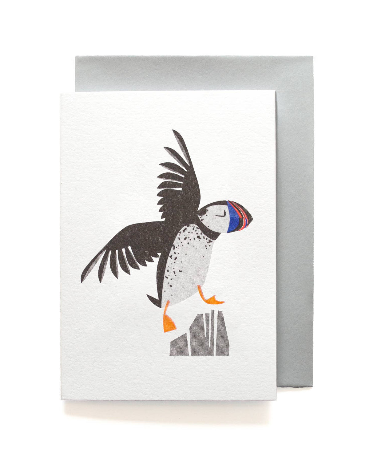 A white textured greeting card featuring a stylized puffin illustration in black, white, gray, orange, and blue, shown with a silver-gray envelope behind it on a white background.