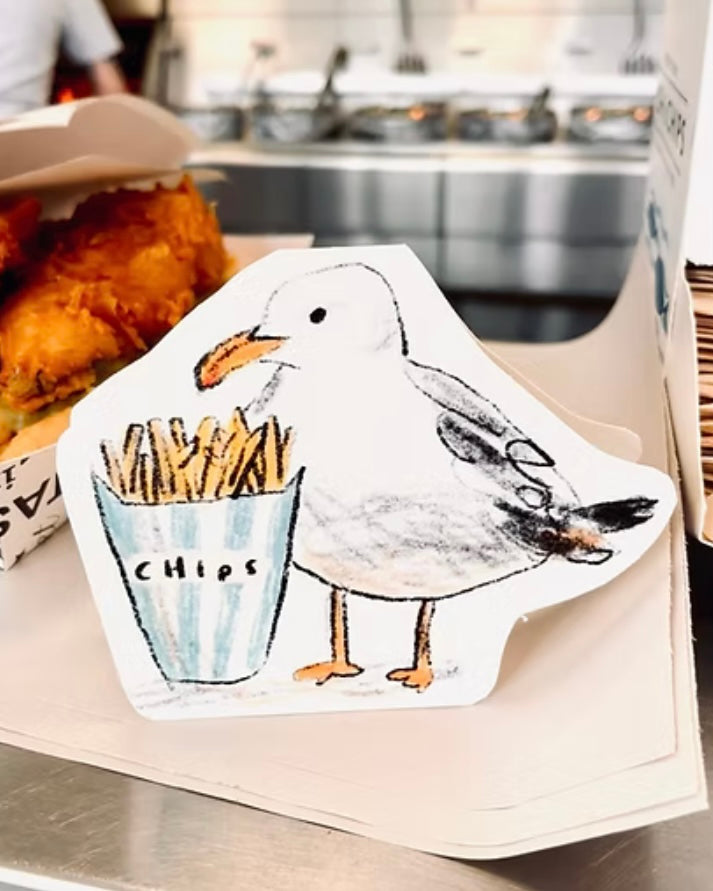 Shaped card with a seagull holding a bucket of chips, on a chip shop counter with fried food in the background.