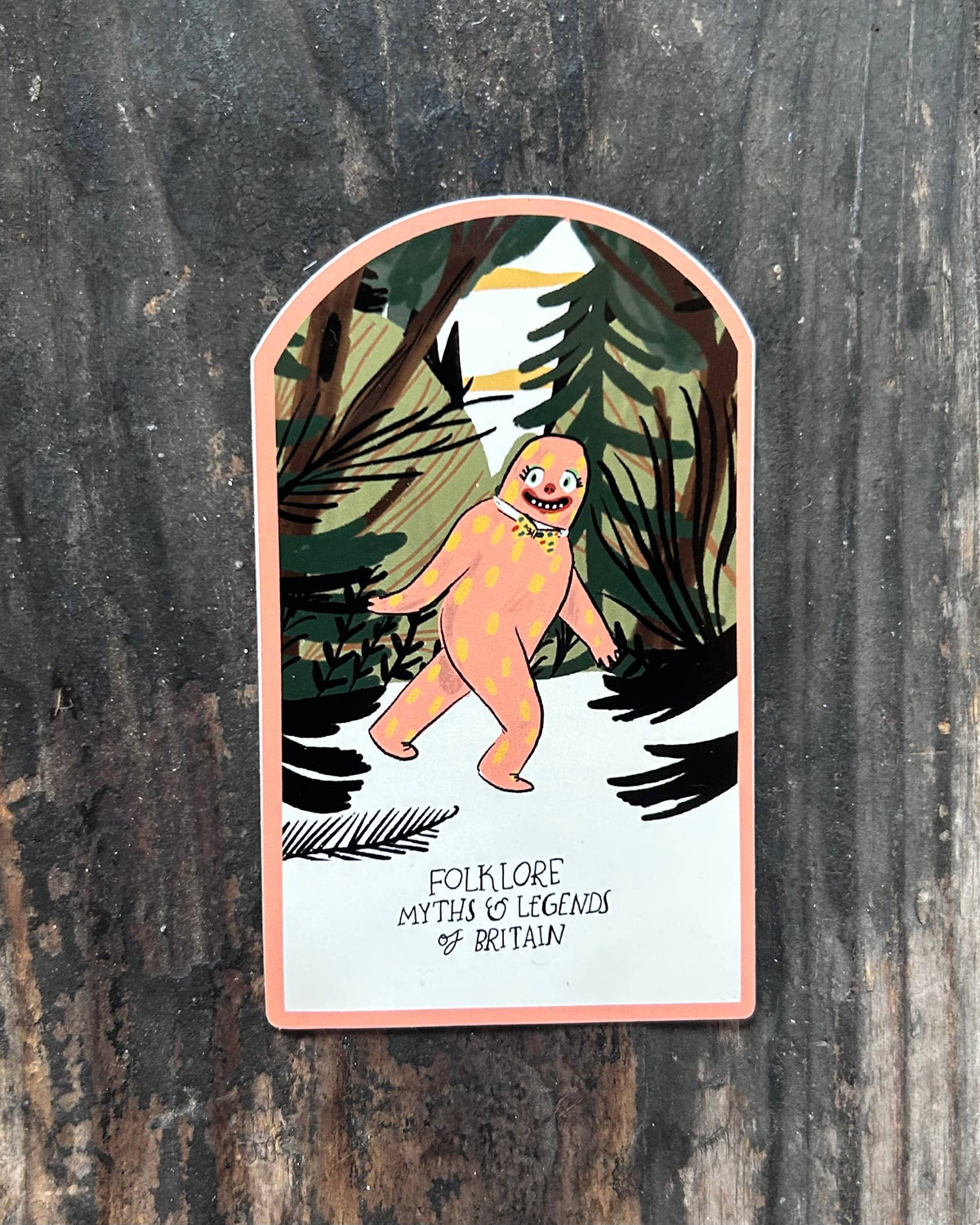 A die-cut sticker featuring an illustration of a pinkish-orange Bigfoot Mr Blobby in a forest, with the text 'FOLKLORE MYTH & LEGENDS OF BRITAIN' at the bottom, shown on a dark weathered wooden background