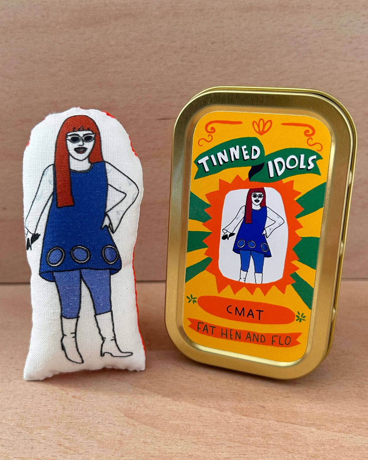 A plush toy featuring a cartoon woman in a blue dress and red hair, placed beside a gold metal tin labeled 'Tinned Idols' on a light wood surface.